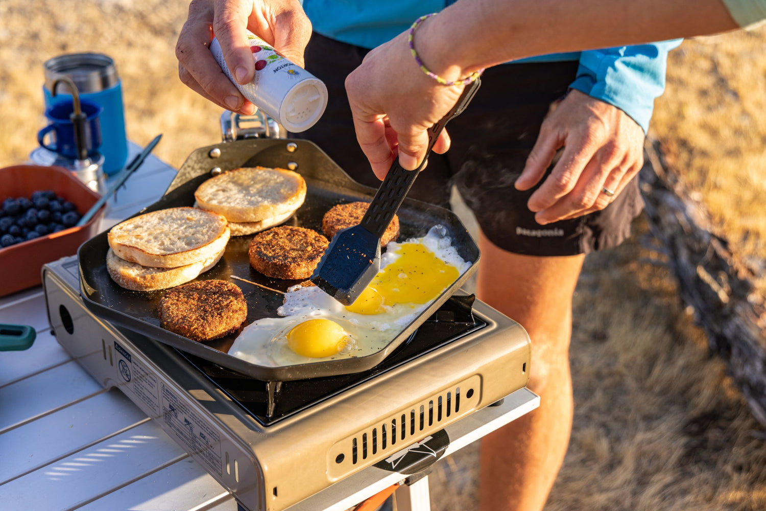 Person cooking breakfast items on a portable stove outdoors using the Compleat Griddle